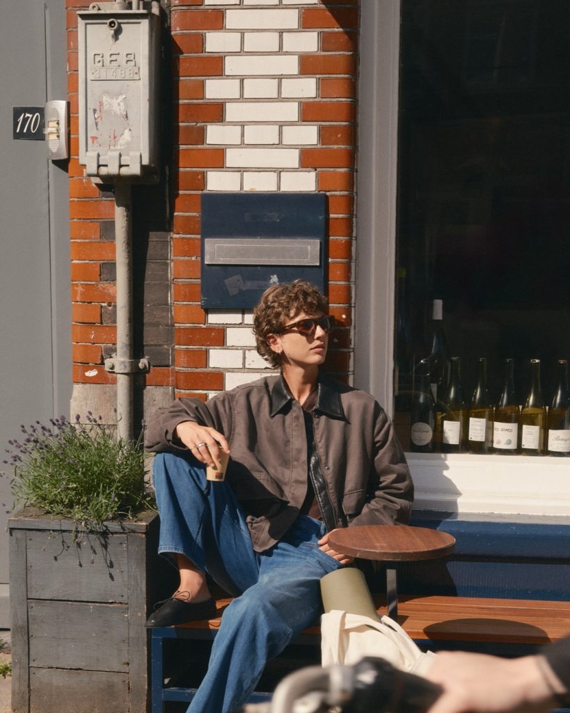 Person in dunklem Hemd und weiter Jeans sitzt in der Sonne vor einer Backsteinfassade