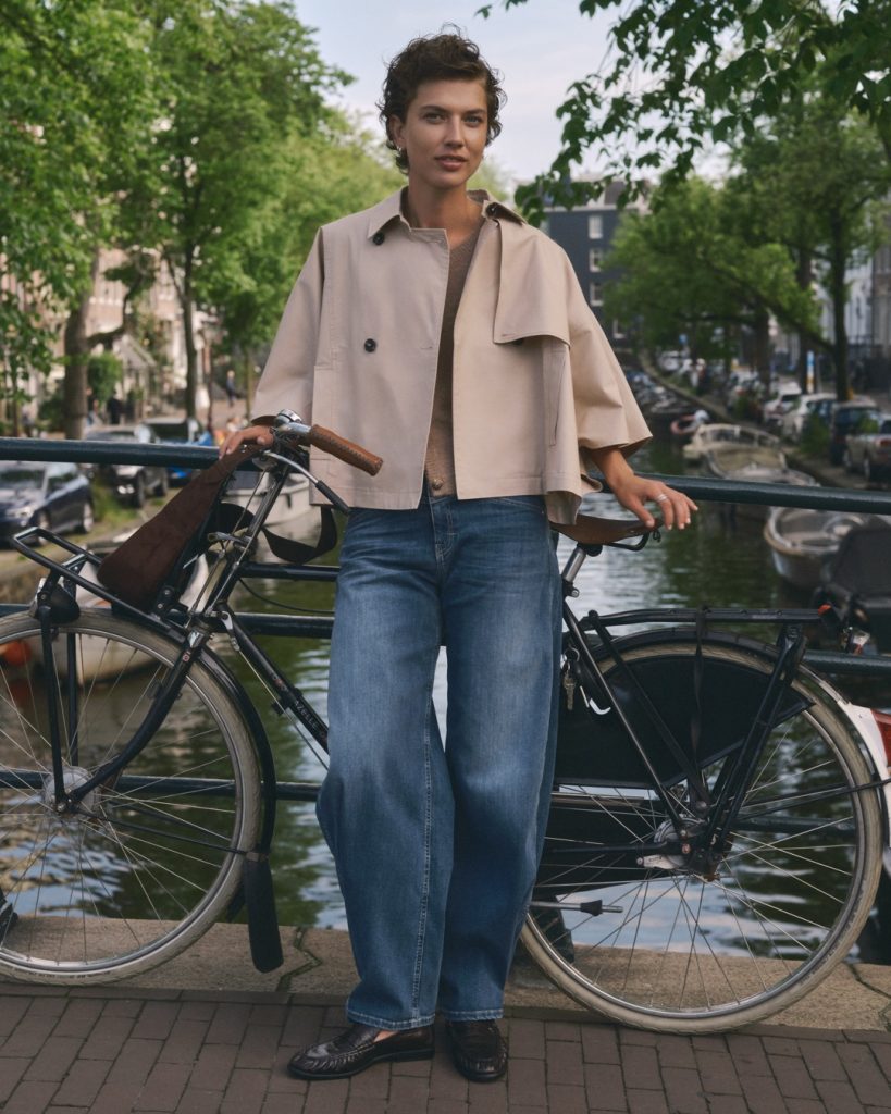 Person in kurzer beiger Jacke und weiter Jeans lehnt an einem Fahrrad an einer Gracht
